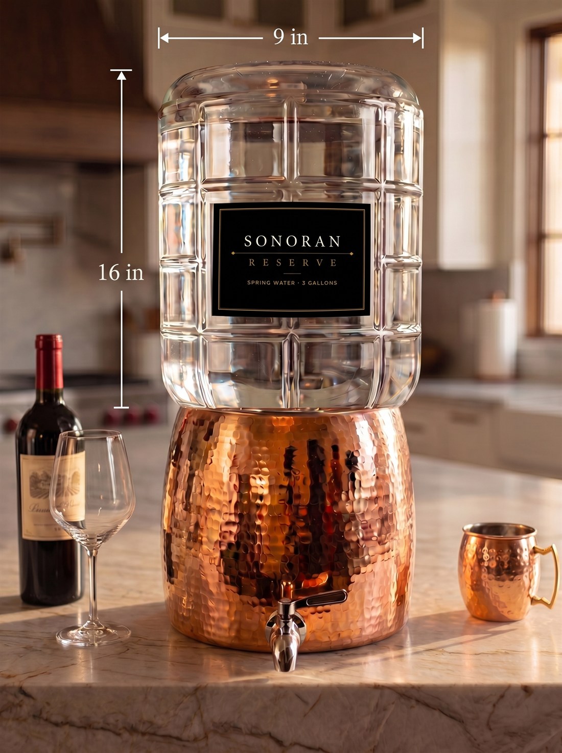 The 3-gallon Sonoran Reserve glass carboy inverted into a hammered-copper dispenser with a stainless-steel spigot, on a marble kitchen counter with a bottle of wine, a wine glass and a copper cup; white floating dimension callouts read 16 inches tall and 9 inches across.